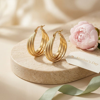 Aramat Jewels gold triple hoop earrings with textured stripe pattern, 25mm diameter, displayed on a stone pedestal with a pink peony and Mother's Day tag.