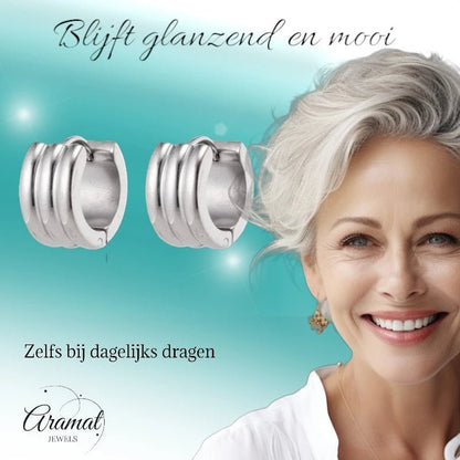 Aramat Jewels wide silver-tone stainless steel hoop earrings with ribbed detail, worn by a smiling woman with grey hair.