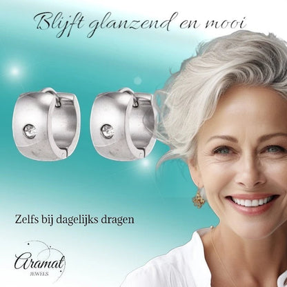 Aramat Jewels silver-colored wide stainless steel hoop earrings with a clear zirconia stone, worn by a smiling woman with grey hair.