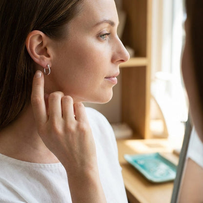 Aramat Jewels silver-tone stainless steel oval hoop earrings worn by a woman with a white shirt.