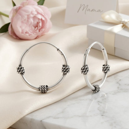 Aramat Jewels thin 16mm silver Bali hoop earrings with a braided detail, presented on a satin cloth with a pink peony and gift box.