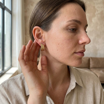 Aramat Jewels goudkleurige triple oorstekers met drie staafjes in een halve hoepel design gedragen door een vrouw.