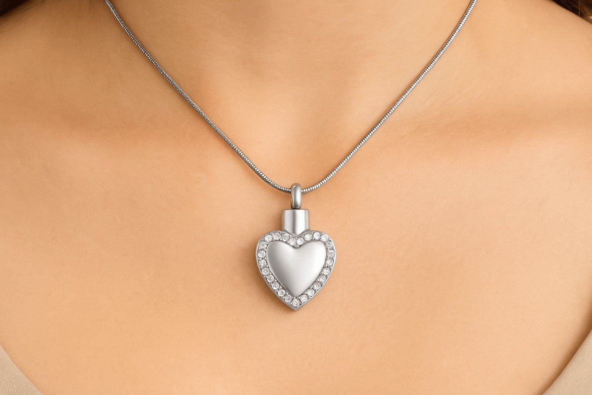 Zilveren hartvormige ashanger met diamanten rand, 33x20mm, gedragen op vrouwenhals met zilveren slangenketting. Luxe gedenksieraad voor asresten, rouwsieraad in hartvorm met kristallen.