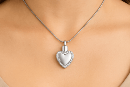Zilveren hartvormige ashanger met diamanten rand, 33x20mm, gedragen op vrouwenhals met zilveren slangenketting. Luxe gedenksieraad voor asresten, rouwsieraad in hartvorm met kristallen.